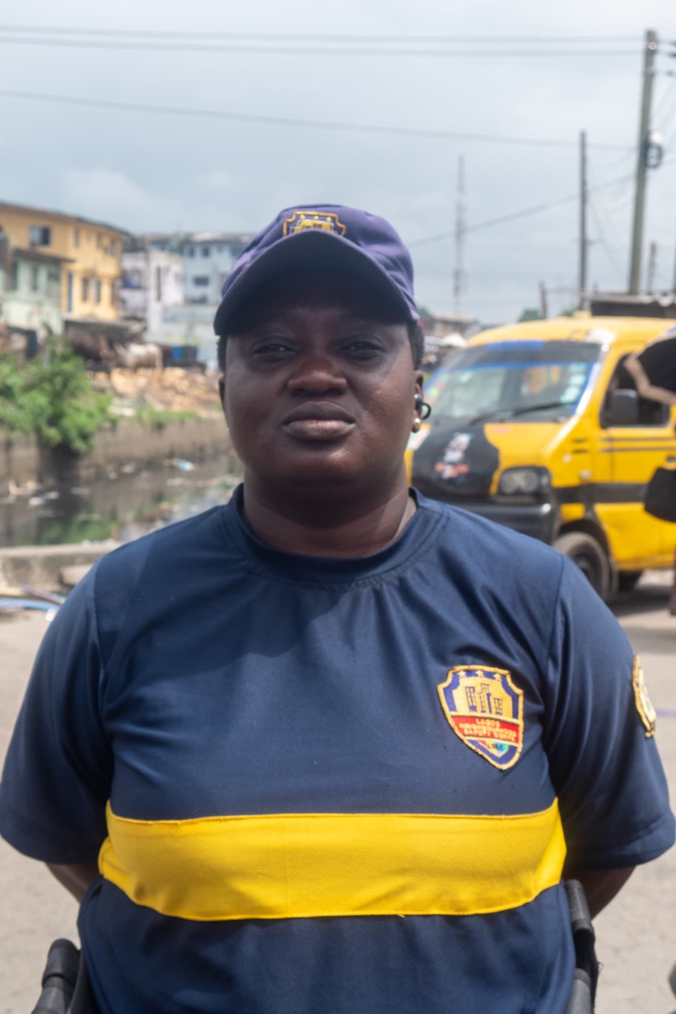 Officer Dada stands in her navy Lagos State Neighbourhood Safety Corps uniform and cap on a Lagos street, a yellow danfo bus behind her.