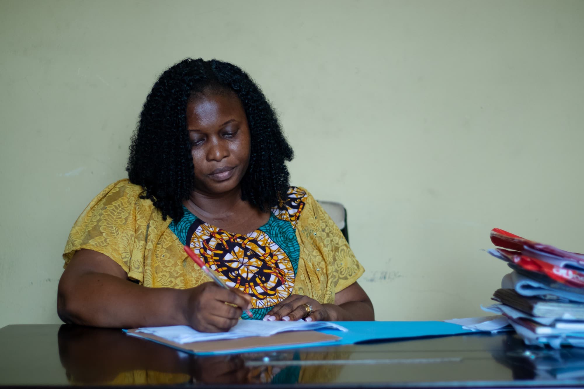 A woman in a yellow lace top over an Ankara print dress writes with a red pen at a desk, a tall stack of files beside her.