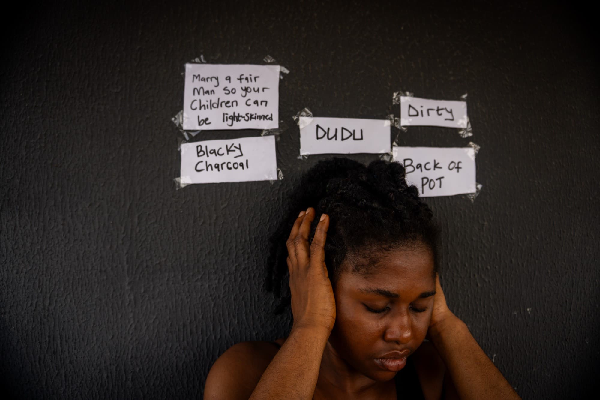 A young woman presses her hands over her ears with her eyes closed, before a dark wall taped with handwritten colourist slurs including “DUDU”, “Dirty”, “Blacky Charcoal”, and “Marry a fair man so your children can be light skinned.”