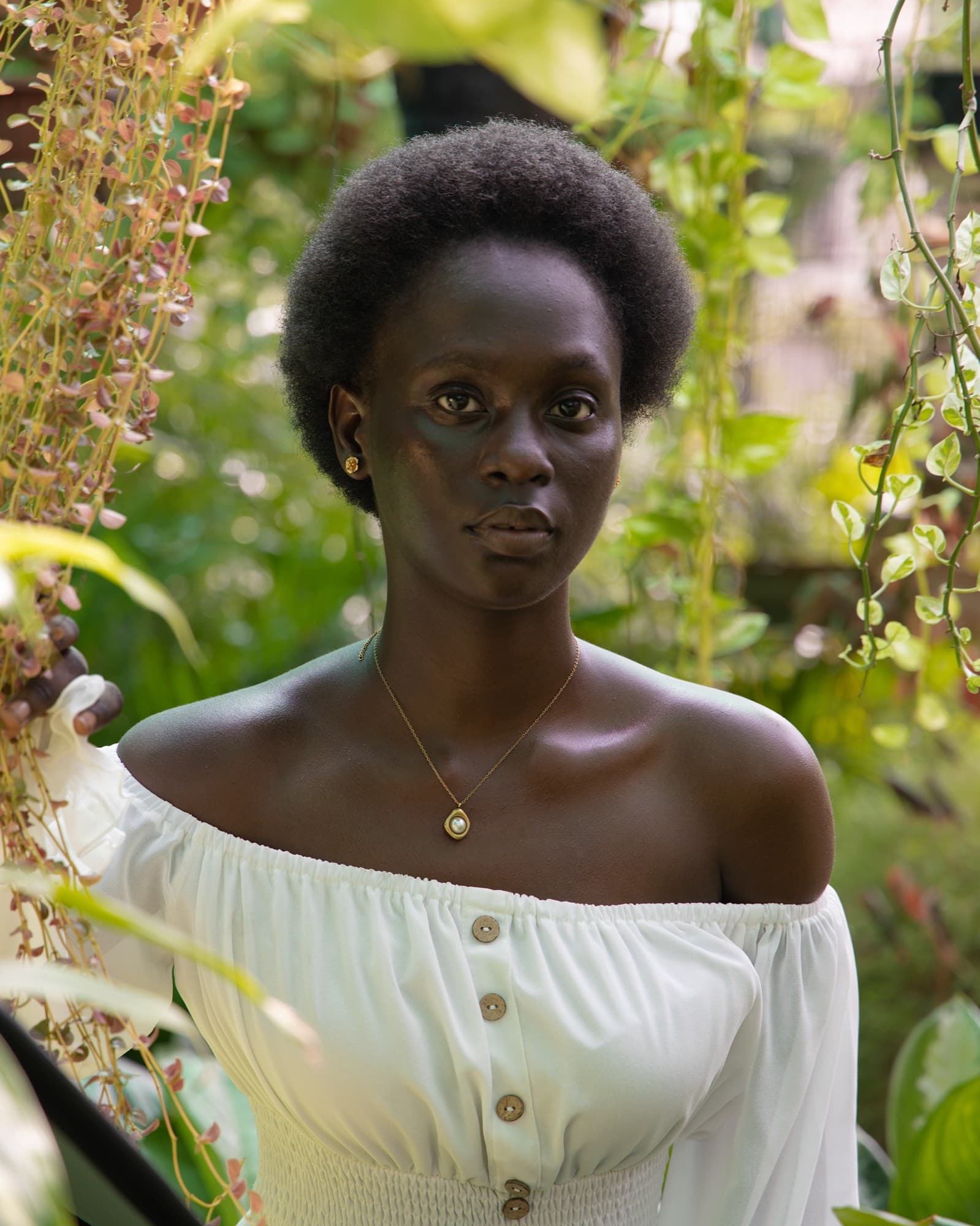 A dark skinned woman with a natural afro, in an off the shoulder white blouse and a small gold pendant, looks directly at the camera amid lush hanging foliage.