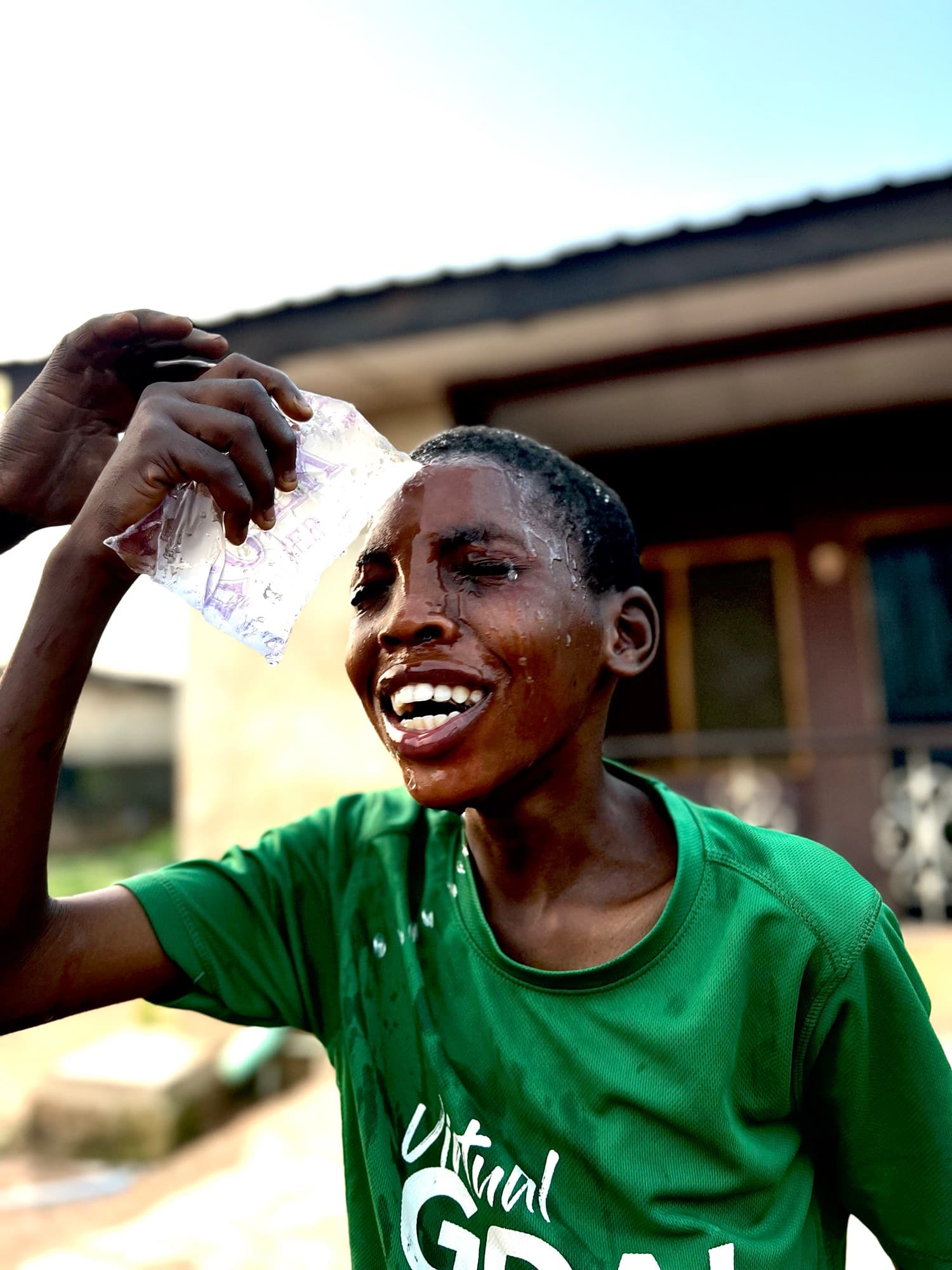 A boy in a green shirt presses a plastic sachet of cold water against his forehead outdoors in bright sun, water streaming down his laughing face, a house with a corrugated metal roof behind him.